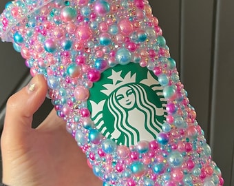 Op de afbeelding: Een doorzichtige herbruikbare beker van plastic bedekt met parelmoer roze, blauwe en witte parels met het Starbucks-logo aan de voorkant.