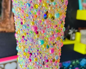Puede incluir: Un vaso de plástico transparente cubierto con una multitud de pedrería y gemas de colores. La taza tiene una tapa blanca y una pajita amarilla. La pedrería es de varios tamaños y colores, creando una superficie brillante y texturizada.