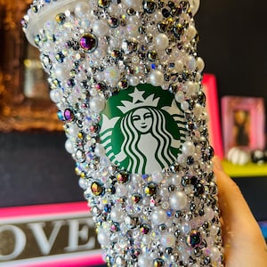 May include: A bedazzled Starbucks tumbler, covered in white pearls, iridescent rhinestones, and iridescent beads. The Starbucks logo is visible in green. The cup is a clear plastic with a white lid.