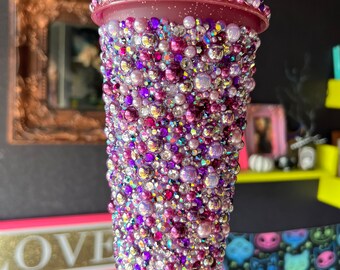 Op de afbeelding: Een paarse herbruikbare beker bedekt met iriserende paarse, roze en witte strass steentjes. De beker heeft een rietje.