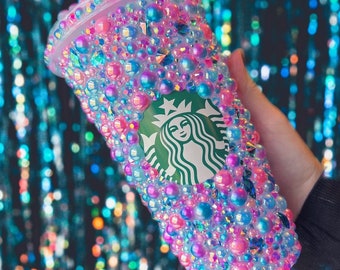 May include: A clear plastic reusable cup covered in iridescent pink, blue, and white gems. The cup has the Starbucks logo on the front.