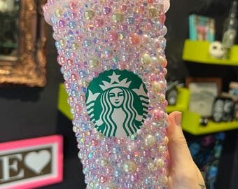 Puede incluir: Un vaso de plástico transparente cubierto de cuentas iridiscentes de color rosa, blanco y amarillo. El vaso tiene el logotipo de Starbucks en la parte delantera.