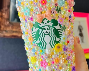 Op de afbeelding: Een versierde Starbucks-beker, versierd met een verscheidenheid aan kleurrijke versieringen. De beker is voorzien van parels, strass-steentjes en bloemaccenten in roze, gele en blauwe tinten. Een perzikstrik en het Starbucks-logo zijn zichtbaar.