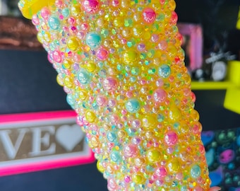 Op de afbeelding: Een gele beker bedekt met iriserende pastelkleurige kralen. De kralen zijn in een willekeurig patroon gerangschikt en creëren een glinsterend effect.