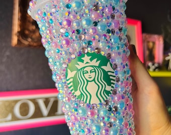 Op de afbeelding: Een doorzichtige plastic beker bedekt met iriserende blauwe, roze en witte strass steentjes. De beker heeft het Starbucks-logo aan de voorkant.