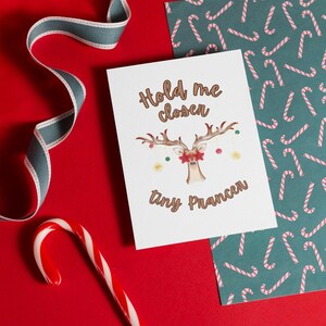 May include: A Christmas card with a reindeer illustration and the text "Hold me closer, tiny prancer." The card is on a red surface with a candy cane, ribbon, and a patterned paper with candy canes.