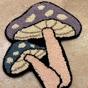 May include: A handmade mushroom-shaped rug with a whimsical design. The rug features a purple mushroom cap with cream-colored spots, a blue mushroom cap, and a pink stem, all outlined in black. The rug is made of a soft, textured material.
