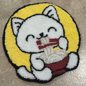 May include: A round, white cat rug with a yellow background and black outline. The cat is smiling and eating ramen from a red bowl. The rug is made of a soft, textured material.