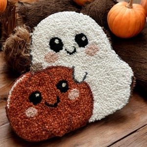 May include: A handmade Halloween-themed rug featuring a white ghost and a brown pumpkin, both with smiling faces and blush accents. The ghost and pumpkin are made with a textured, looped yarn. The rug is likely intended for seasonal home decor.