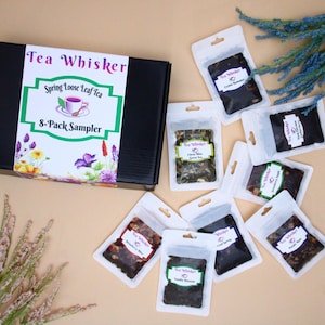 May include: A black box with the text "Tea Whisker Spring Loose Leaf Tea 8-Pack Sampler" on the front. The box is surrounded by eight small clear plastic bags filled with loose leaf tea. Each bag has a label with the text "Tea Whisker" and the type of tea.
