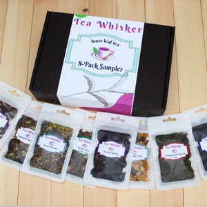 May include: A black box with a white label that says "Tea Whisker loose leaf tea 8-Pack Sampler". The box contains eight small clear bags of loose leaf tea with different labels. The labels are white with black text and feature different tea names, such as "Cinnamon Roll" and "Sweet Dreams".