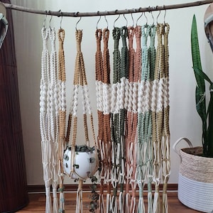Könnte beinhalten: Eine Sammlung von Makramee-Pflanzenhängern in verschiedenen Farben, darunter Beige, Braun, Grün und Orange. Die Hänger sind aus Baumwollschnur gefertigt und haben ein geknüpftes Design. Eine kleine Topfpflanze hängt an einem der Hänger.