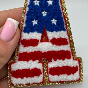 May include: A letter "A" patch with a gold glitter border. The patch features the American flag design, with blue stars on a blue background and red and white stripes. The patch is made of a textured material.