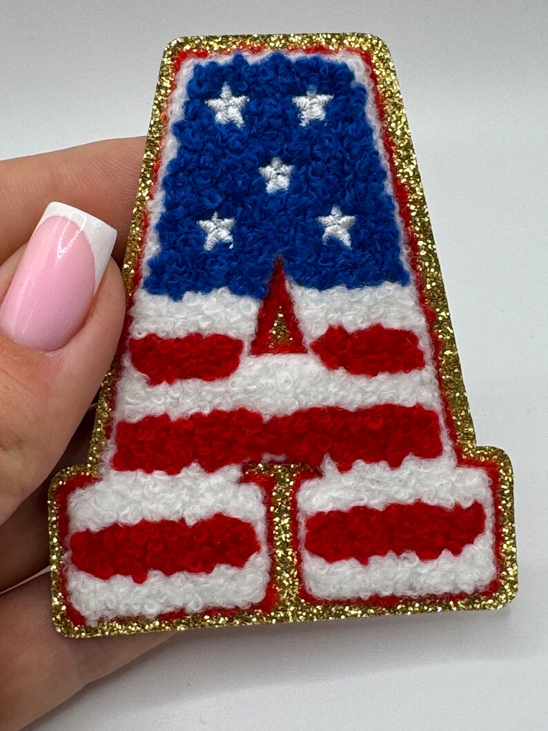 May include: A letter "A" patch with a gold glitter border. The patch features the American flag design, with blue stars on a blue background and red and white stripes. The patch is made of a textured material.