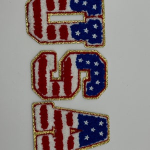May include: Three embroidered patches spelling "USA". Each letter is white with red stripes, blue star accents, and a gold glitter border. The patches are likely for clothing or accessories.