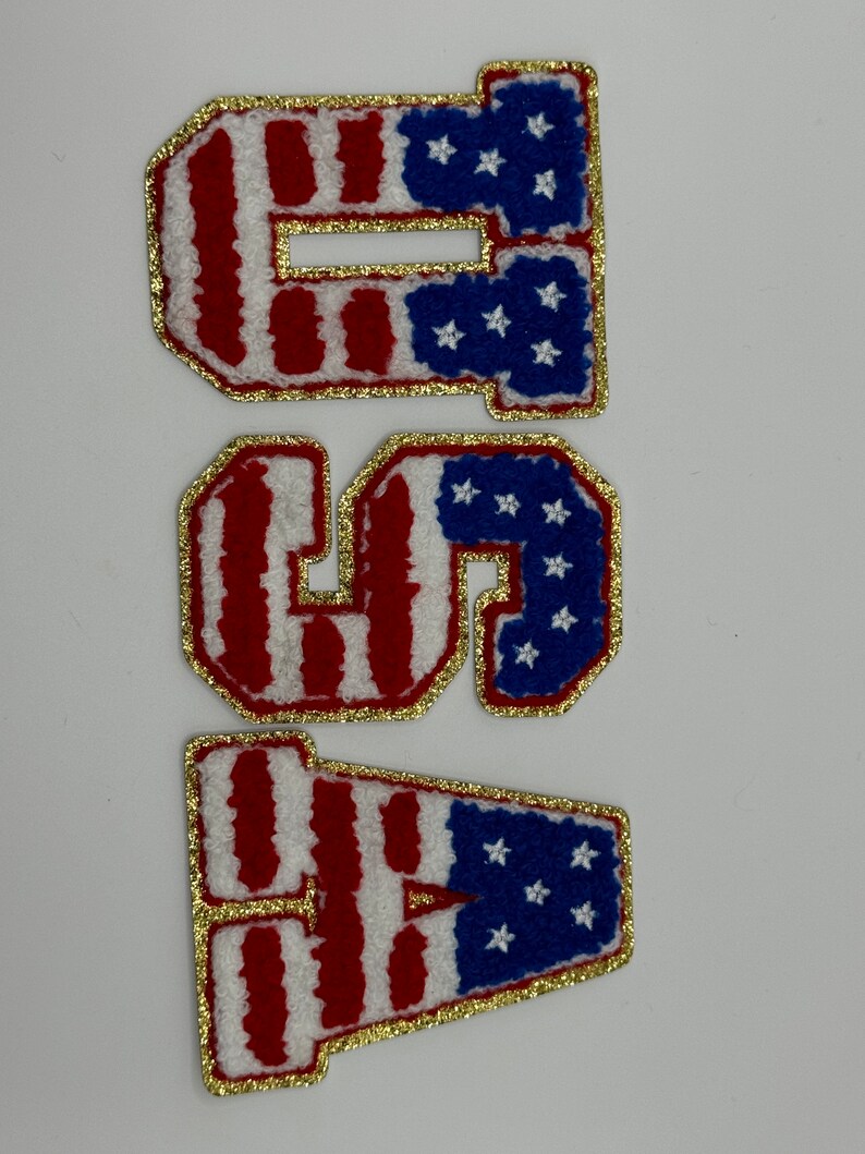 May include: Three embroidered patches spelling "USA". Each letter is white with red stripes, blue star accents, and a gold glitter border. The patches are likely for clothing or accessories.