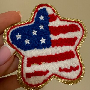 May include: A star-shaped patch featuring the American flag design. The patch has a blue field with white stars and red and white stripes. The edges are outlined with gold glitter.