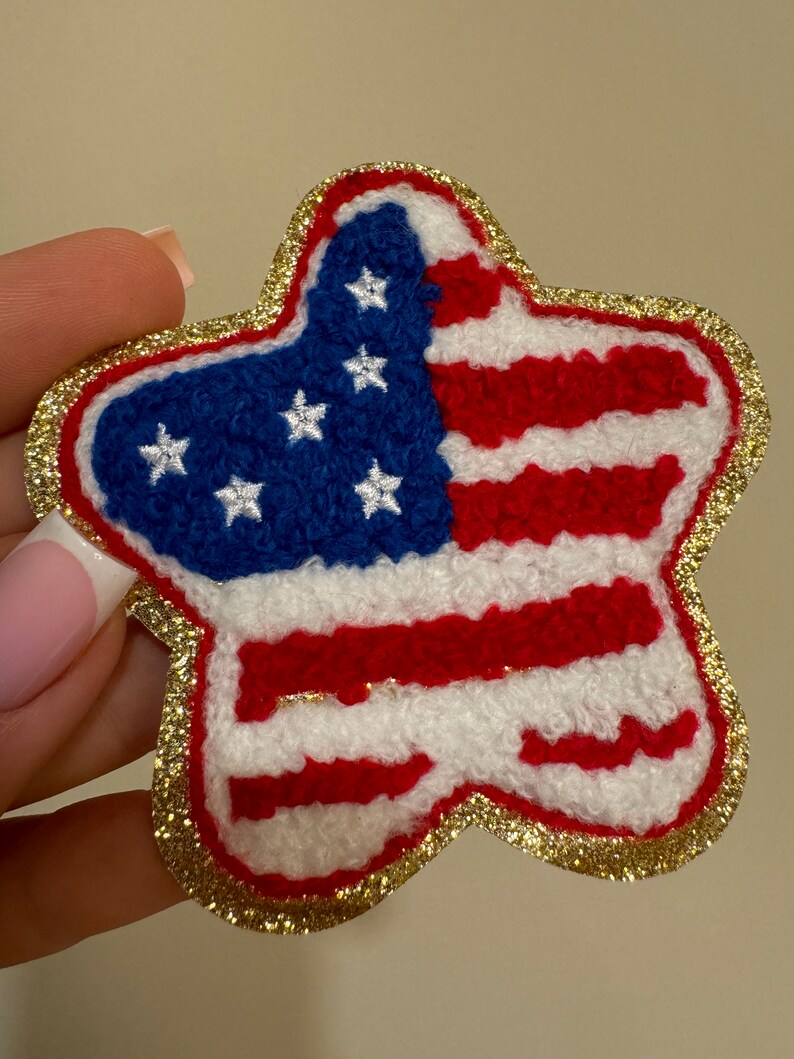 May include: A star-shaped patch featuring the American flag design. The patch has a blue field with white stars and red and white stripes. The edges are outlined with gold glitter.