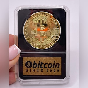 May include: A gold-colored Bitcoin coin in a clear plastic case with a black background. The coin has the Bitcoin symbol and the text "Digital Decentralized Peer to Peer" around the edge. The case also has a gold-colored label with the text "bitcoin SINCE 2009".