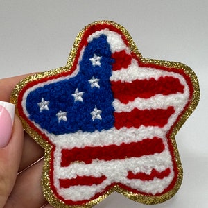 May include: A star-shaped patch featuring the American flag design. The patch has a blue field with white stars and red and white stripes. It is outlined with gold glitter.