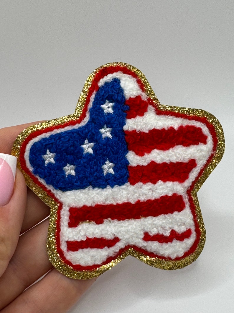 May include: A star-shaped patch featuring the American flag design. The patch has a blue field with white stars and red and white stripes. It is outlined with gold glitter.