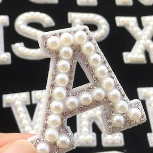 May include: A capital letter A patch featuring a white felt base adorned with a border of small clear rhinestones and a central inlay of round white faux pearls. The letter is designed for attachment to fabric surfaces as a decorative element.