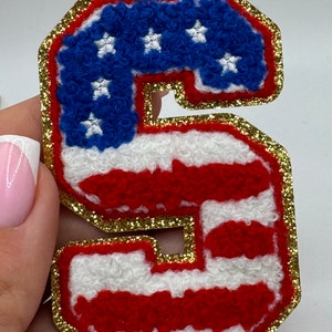 May include: A patriotic letter "S" patch with a gold glitter border. The patch features a blue field with white stars, and red and white stripes, resembling the American flag. This decorative patch is ideal for apparel and accessories.