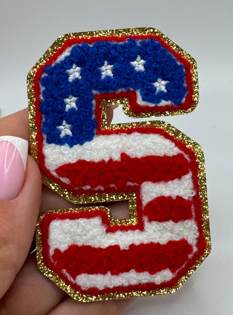 May include: A patriotic letter "S" patch with a gold glitter border. The patch features a blue field with white stars, and red and white stripes, resembling the American flag. This decorative patch is ideal for apparel and accessories.