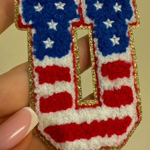 May include: A patriotic letter "U" patch with a gold glitter border. The letter features a design of the American flag, with red and white stripes and a blue field with white stars. The patch is likely for clothing or accessories.