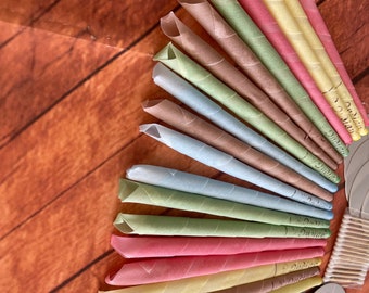 May include: A variety pack of ear candles in a rainbow of colors, including blue, green, yellow, pink, and brown. The candles are arranged in a fan shape on a wooden surface.