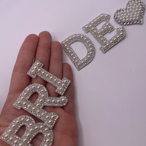 May include: A set of pearl-embellished letters spelling "BRIDE" and a pearl heart. The letters are white with a silver backing and are made of small, white pearls.