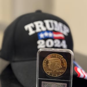 May include: A gold-colored coin with the words "Never Surrender" and a portrait of Donald Trump. The coin is in a clear plastic case with a black background. The coin also has the words "In God We Trust" and "2024".