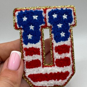 May include: A patriotic letter "U" patch, featuring the American flag design. The letter is in red, white, and blue, with a gold glitter border. The stars are white, and the stripes are red and white.