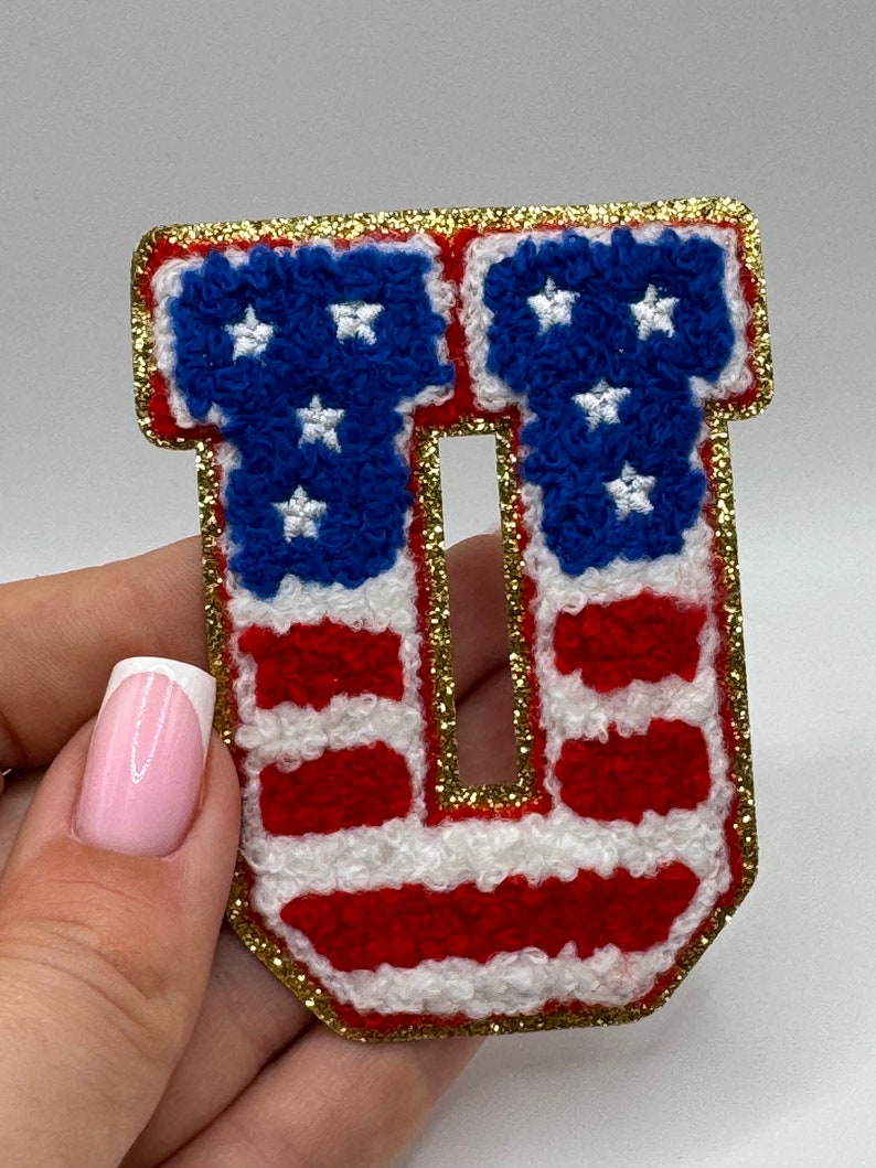 May include: A patriotic letter "U" patch, featuring the American flag design. The letter is in red, white, and blue, with a gold glitter border. The stars are white, and the stripes are red and white.