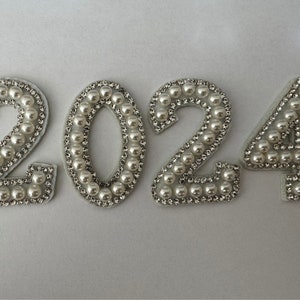 Peut inclure: Un ensemble de quatre chiffres en tissu blanc, 2024, décorés de perles blanches et de strass argentés.