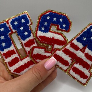 May include: Embroidered patches spelling out "USA" in red, white, and blue, resembling the American flag. The letters have a gold glitter border. The patches are likely for clothing or accessories.