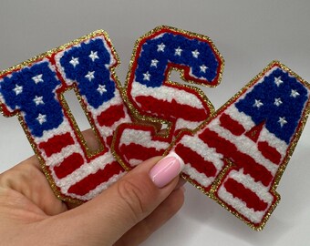 May include: Embroidered patches spelling out "USA" in red, white, and blue, resembling the American flag. The letters have a gold glitter border. The patches are likely for clothing or accessories.