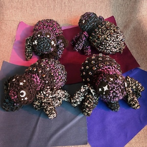 May include: Four crocheted spider plushies in various shades of purple, pink, and black with white accents. Each spider has a beaded body and legs.