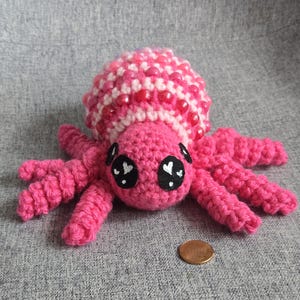 May include: A pink crocheted spider with white and pink beads around its body and black eyes with white hearts. The spider is sitting on a gray surface.