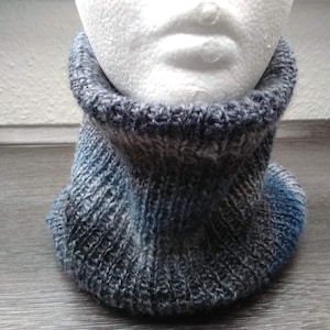 Könnte beinhalten: Ein gestrickter Rundschal in Blau-, Grau- und Weißtönen. Der Schal ist aus weichem, strukturiertem Garn gefertigt und hat ein Rippenmuster. Er wird auf einem weißen Schaufensterkopf präsentiert. Dieser Halswärmer ist perfekt für kaltes Wetter.