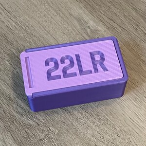 Peut inclure: Une bo&icirc;te en plastique violette avec un couvercle coulissant et le texte "22LR" imprim&eacute; sur le dessus.