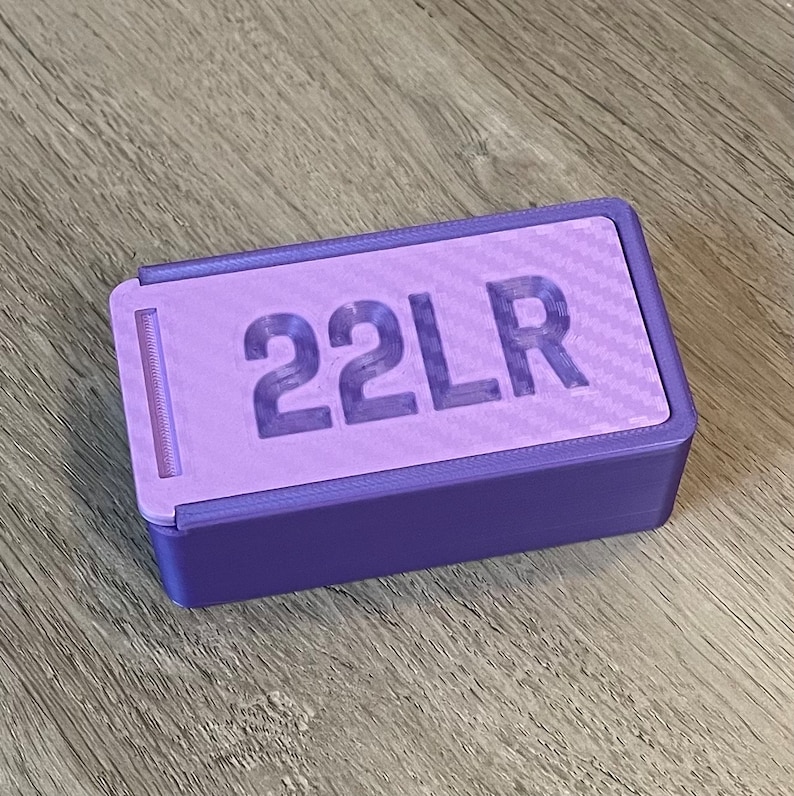 Peut inclure: Une bo&icirc;te en plastique violette avec un couvercle coulissant et le texte "22LR" imprim&eacute; sur le dessus.