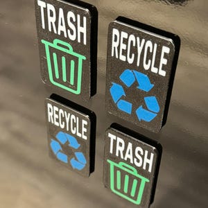 Peut inclure: Quatre aimants carrés noirs avec du texte blanc et des icônes colorées. Deux aimants disent "TRASH" avec une icône de poubelle verte. Deux aimants disent "RECYCLE" avec un symbole de recyclage bleu.