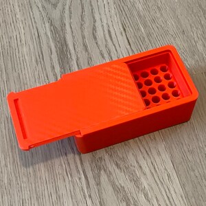 Peut inclure: Une bo&icirc;te en plastique orange vif avec un couvercle &agrave; charni&egrave;re et une grille de trous ronds &agrave; l'int&eacute;rieur. La bo&icirc;te est con&ccedil;ue pour contenir de petits objets, tels que des balles ou des perles.