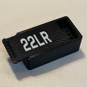 Peut inclure: Une bo&icirc;te en plastique noire avec un couvercle &agrave; charni&egrave;re et une &eacute;tiquette blanche indiquant "22LR". La bo&icirc;te pr&eacute;sente un motif en fibre de carbone sur le couvercle et une rang&eacute;e de trous &agrave; l'int&eacute;rieur.