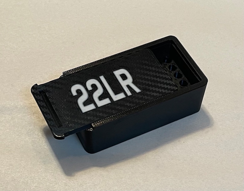 Peut inclure: Une bo&icirc;te en plastique noire avec un couvercle &agrave; charni&egrave;re et une &eacute;tiquette blanche indiquant "22LR". La bo&icirc;te pr&eacute;sente un motif en fibre de carbone sur le couvercle et une rang&eacute;e de trous &agrave; l'int&eacute;rieur.