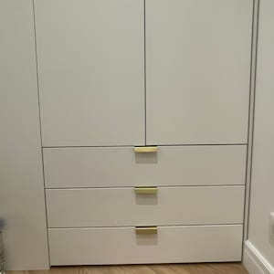 Puede incluir: Armario blanco con tres cajones y tiradores de color dorado. El armario está cerrado y los cajones están cerrados.