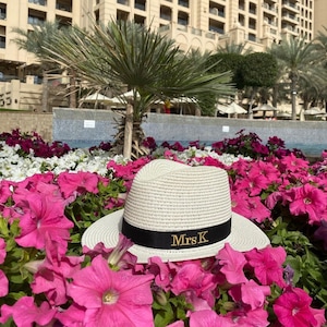 May include: A cream-coloured fedora hat with a black band and gold embroidered text "Mrs K" sits amongst vibrant pink flowers. The hat is made of woven material and has a classic brim.
