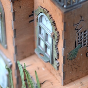 May include: A detailed wooden model of a haunted house with intricate laser-cut details. The house features windows with green trim, a cracked wall, and a green hand reaching out from a barred window. The model is made of light brown wood.