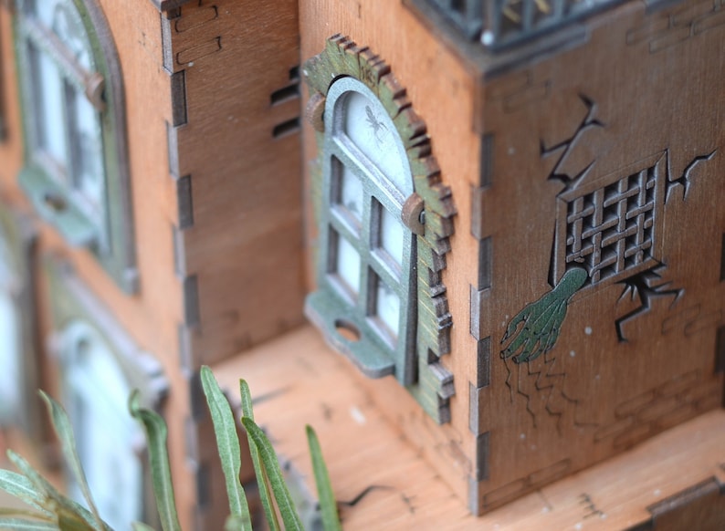 May include: A detailed wooden model of a haunted house with intricate laser-cut details. The house features windows with green trim, a cracked wall, and a green hand reaching out from a barred window. The model is made of light brown wood.